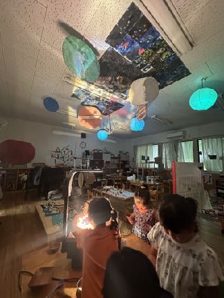 天井に飾った惑星や星空に光を当てた保育室で遊ぶ子どもたち